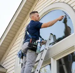 Professional window cleaner on a ladder using a squeegee to clean an exterior arched window, Birmingham, AL.